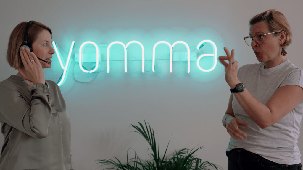 Zwei Mitarbeiterinnen von der yomma GmbH im Studio und Berlin. Sie zeigen eine Situation Dolmetschen, Beraten, Textredaktion für BITV 2.0 Webseitentexten