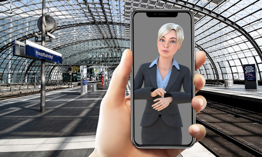 Auf einem Smartphone Handydisplay ist der AVASAG Gebärdensprach-Avatar abgebildet aus dem Verbund- und Forschungsprojekt an dem yomma beteiligt ist. Im Hintergrund der Hauptbahnhof Berlin