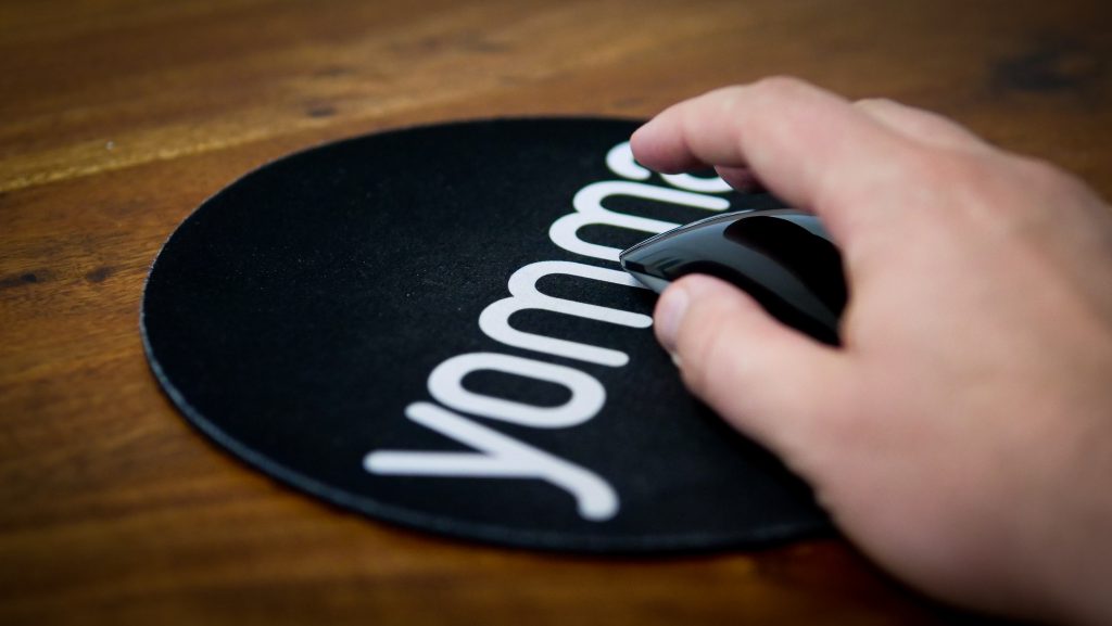 Hand am Computer auf einem mousepad mit dem yomma logo weiß auf schwarz. yomma steht für Gebärdensprache, Gebärdensprache lernen und Gehörlosenkommunikation. Die Maus wird gerade für die Arbeit an einem Projekt wie Gebärdensprachvideos, SQAT oder BITV 2.0 benutz.