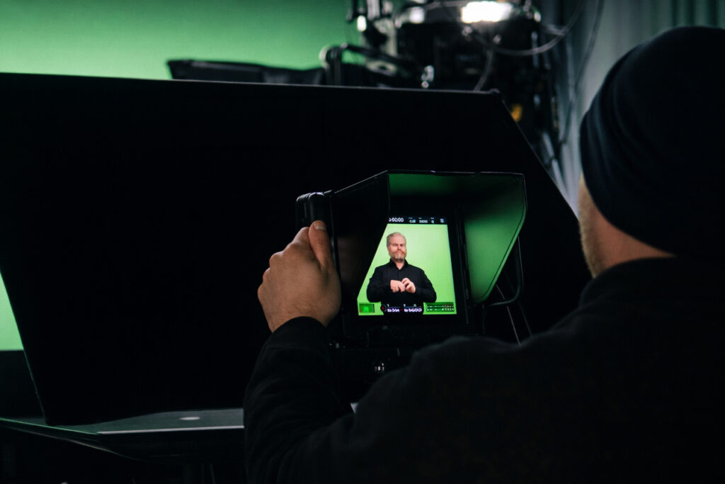 Ein Kameramann nimmt in einem Studio ein Gebärdensprachvideo auf: Eine Person steht vor einem grünen Hintergrund (Greenscreen) und verwendet Gebärdensprache. Auf dem Kameramonitor ist die aufnehmende Person sichtbar, während Kameraequipment und Studiobeleuchtung im Vordergrund zu sehen sind.