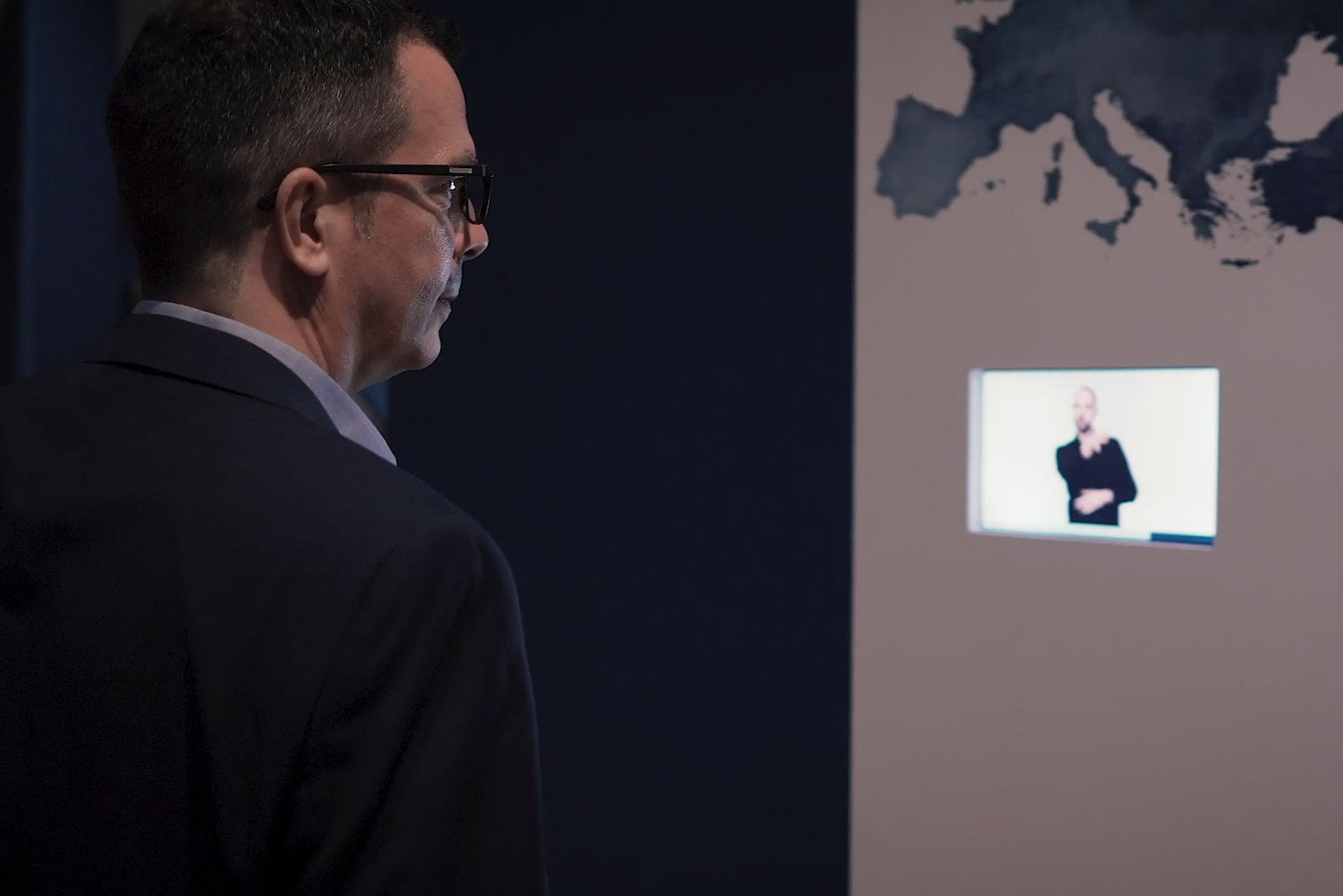 Ein Mann betrachtet eine Präsentationsfläche, auf der ein kleines eingeblendetes Gebärdensprachvideo zu sehen ist. Die Gebärdensprachdolmetscherin übersetzt Inhalte in Deutscher Gebärdensprache (DGS) und macht Präsentationen, Vorträge oder digitale Informationsangebote barrierefrei und für gehörlose Menschen zugänglich.